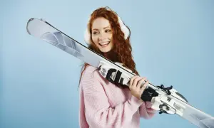 Uśmiechnięta rudowłosa dziewczyna w różowym swetrze trzyma narty. Dowiedz się, jak ustawić siłę wypięcia wiązań narciarskich dla bezpieczeństwa.