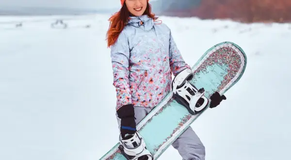 Uśmiechnięta dziewczyna w czapce i kurtce z kwiatowym wzorem trzyma **co to jest snowboard** z kolorowymi zdobieniami.