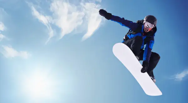 Snowboardzistka w locie na tle błękitnego nieba. Jej deska snowboardowa damska jest biała i lśniąca.