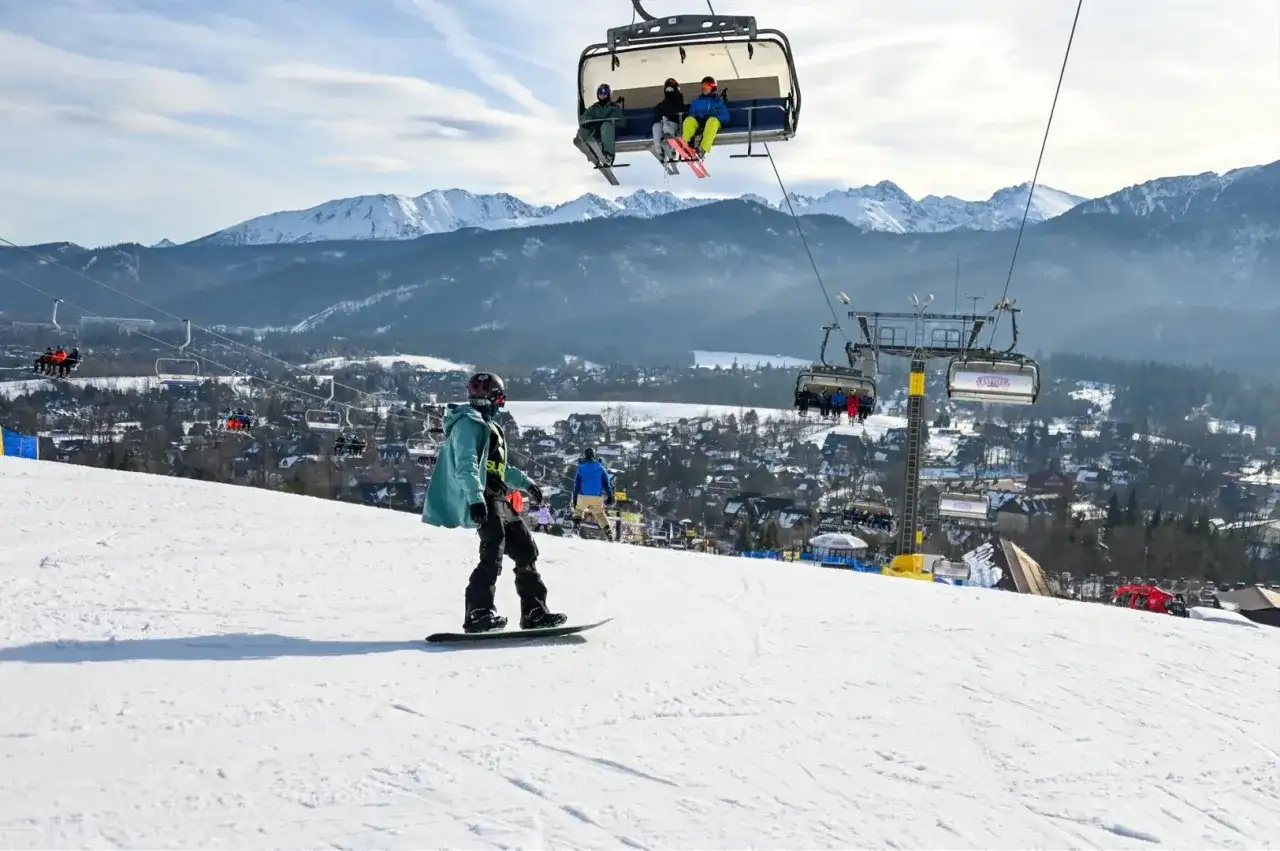Snowboardzista na stoku, w tle kolejka krzesełkowa i góry. Zastanawiasz się, ile kosztuje karnet na narty?