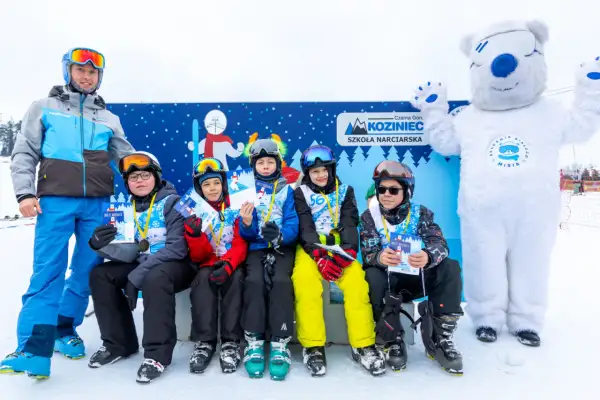 Uśmiechnięci młodzi narciarze z dyplomami i medalami po zajęciach w szkole narciarskiej Koziniec. Towarzyszy im instruktor i biały miś.